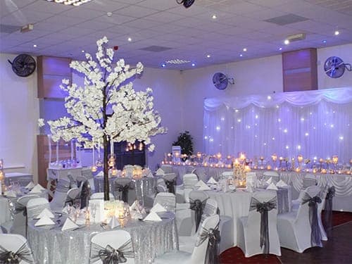 Function room with a large white blossom tree centrepiece, round tables dressed in silver sequin tablecloths and fairy light curtain backdrop